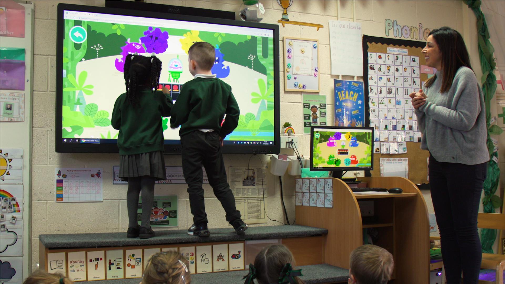 Children playing Number Skills on the interactive whiteboard in a classroom. A teacher watches them.